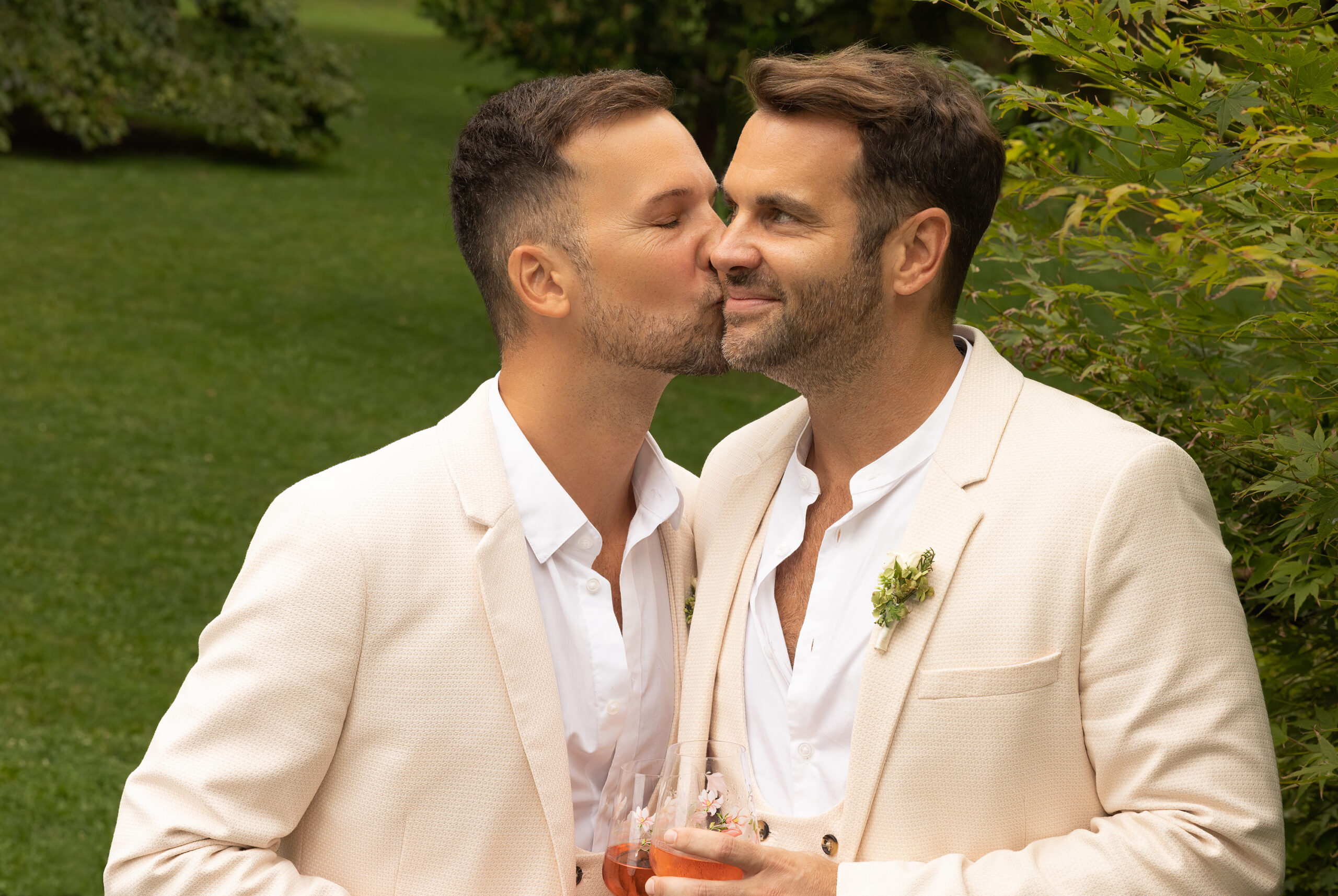 Zwei Männer in Hochzeitsanzügen in einem Park. Der eine Mann küsst den anderen auf die Wange.